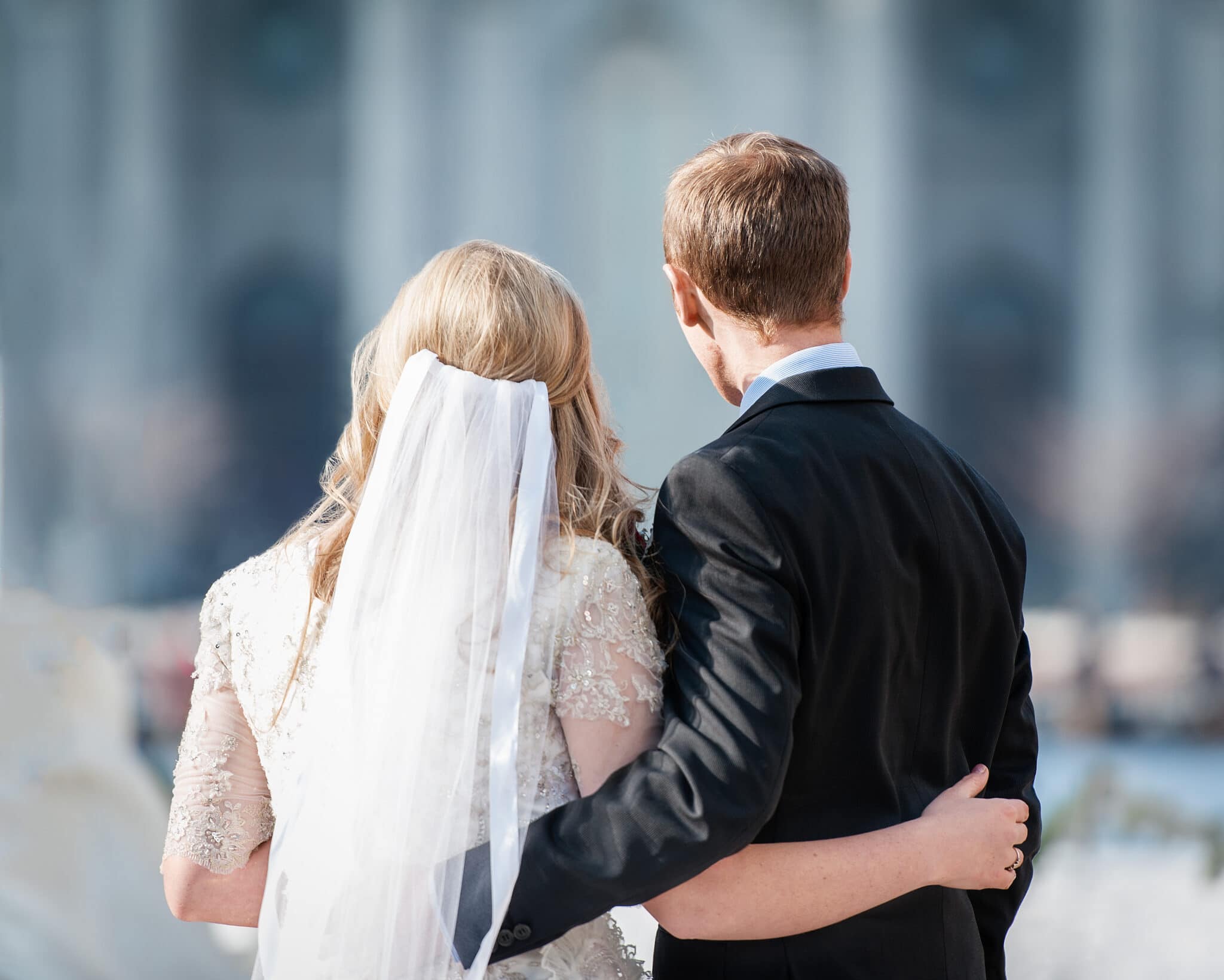 A backside view of a newly married couple that are holding their arms around each other as they stop in front of an LDS temple blurred in the background.
