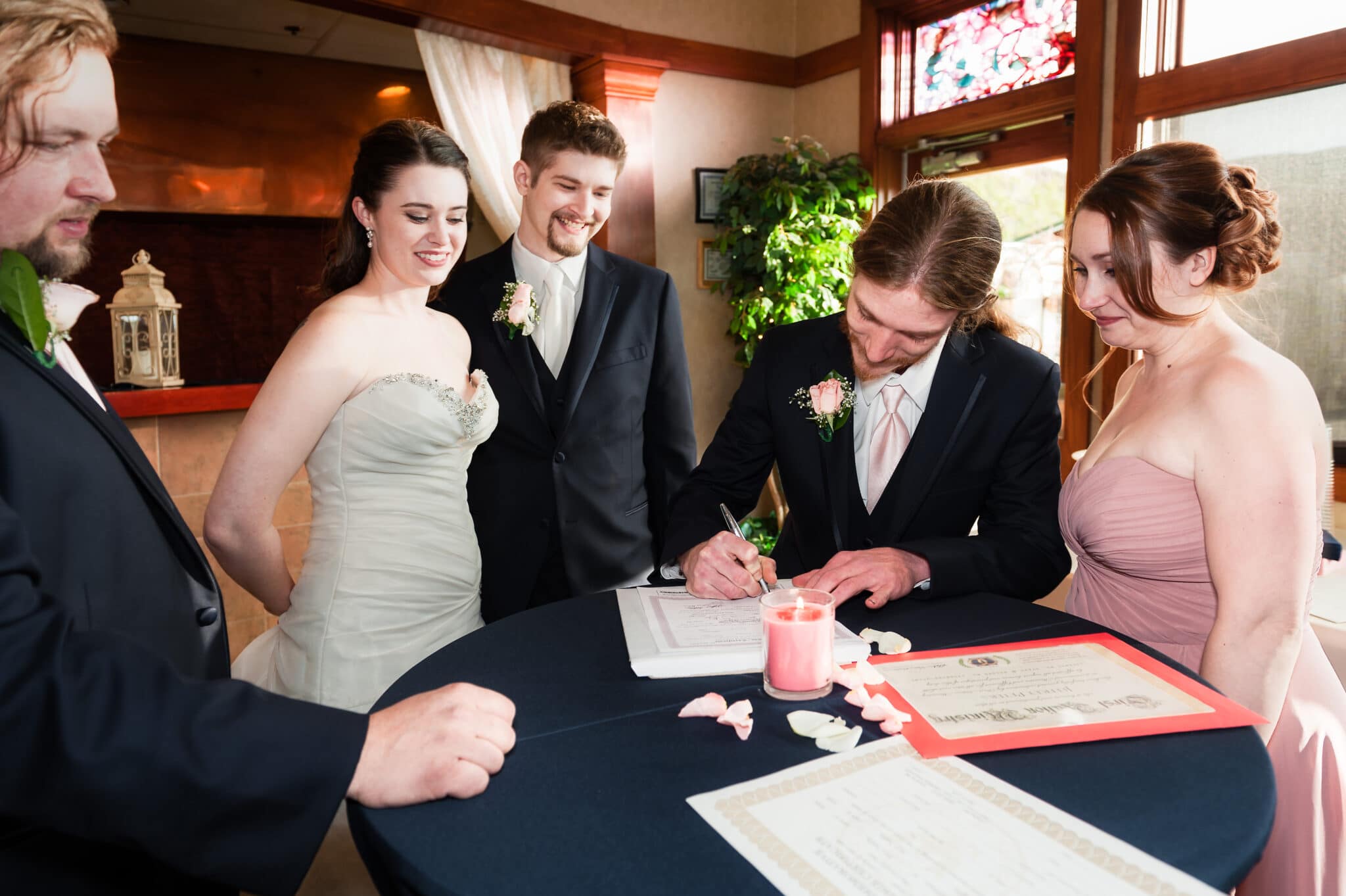 A recently married couple, the officiant and the matron of honor watch as the best man signs his name to witness the event on the marriage license.